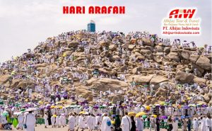 Pilgrims in white robes crowd a rocky hill for Hari Arafah, many umbrellas, travel branding in the corner.