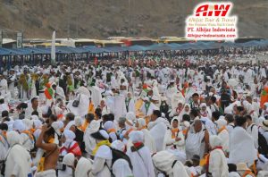 Large crowd of pilgrims in white robes gathered outdoors, with a travel agency logo in the corner (Umrah/Hajj context).