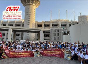 Group of pilgrims posing with red banners that read Jamaah Haji Khusus PT. Alhijaz Indonesia, in front of a modern building with a tower and the Alhijaz logo in the corner logo area.