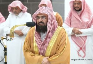 Man in traditional golden robe and red-and-white checkered keffiyeh speaks into a microphone at a formal gathering, with other similarly dressed men in the background.
