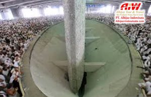 Crowd of pilgrims encircling a circular open shaft with a tall central column inside a mosque; travel company logo in the corner.